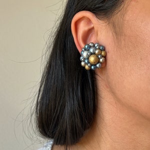 May include: A cluster earring featuring a mix of gray, gold, and white faux pearls. The earring is round and has a textured appearance, with a central gold-colored pearl surrounded by smaller pearls.