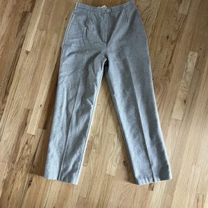May include: A pair of light gray high-waisted trousers. The pants have a button closure and pleats at the waist. The fabric appears to be a wool blend. The pants are laid flat on a wooden surface.