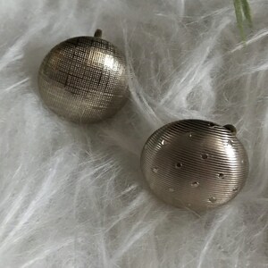 May include: Two silver-toned, round earrings. One earring has a crosshatch pattern, while the other features horizontal lines and small, evenly spaced holes. Both have a small loop at the top for attachment.