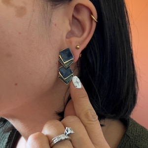 May include: Geometric drop earrings with a layered square design. The earrings feature a dark blue enamel finish with gold-colored edges. The earrings are worn on a person's ear.