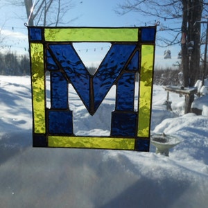 May include: Stained glass panel featuring a blue and yellow letter "M" design. The square panel is framed in yellow and the letter is blue. The panel is hanging in front of a snowy outdoor scene.
