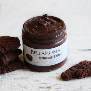 May include: A glass jar of dark brown brownie batter facial butter with the label "BELLAROMA Brownie Batter Gentle Facial Butter raw cacao powder rich cocoa walnut shells 2 oz"