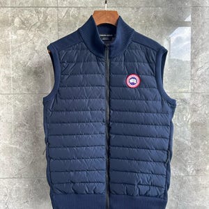 May include: A navy blue quilted vest featuring a full-length front zipper, a ribbed stand-up collar, and ribbed hem. The garment includes a circular Canada Goose patch on the left chest and side panels with a textured knit pattern.