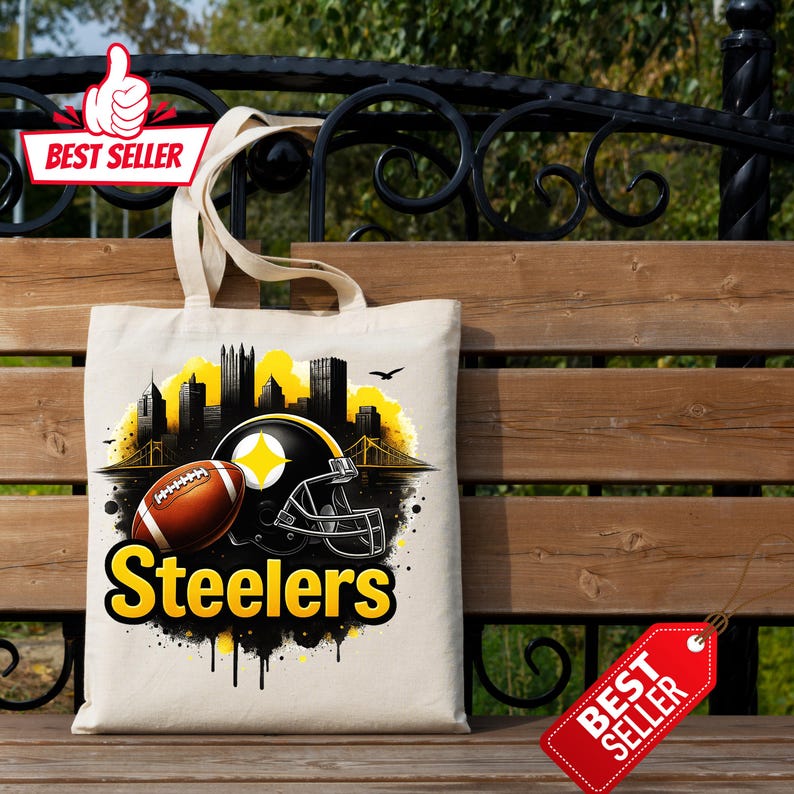 K&ouml;nnte beinhalten: Beige Tragetasche mit einem Football, Helm und dem Wort "Steelers" in Gelb. Das Design beinhaltet eine Stadtlandschaft in Schwarz und Gelb, mit einer Br&uuml;cke und V&ouml;geln. Die Tasche hat ein "Bestseller"-Etikett.