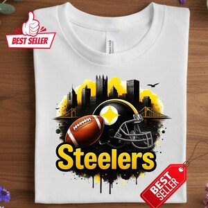 K&ouml;nnte beinhalten: Wei&szlig;es T-Shirt mit einem gelb-schwarzen Design eines Footballs, Helms und einer Skyline. Das Wort "Steelers" ist gelb. Ein "Bestseller"-Etikett ist angebracht.