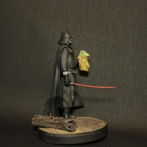 May include: A Star Wars action figure featuring Darth Vader holding a small Grogu figure and a red lightsaber. The figures are on a round base with a piece of wood. The figures are black, green, and tan.