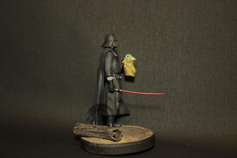 May include: A Star Wars action figure featuring Darth Vader holding a small Grogu figure and a red lightsaber. The figures are on a round base with a piece of wood. The figures are black, green, and tan.