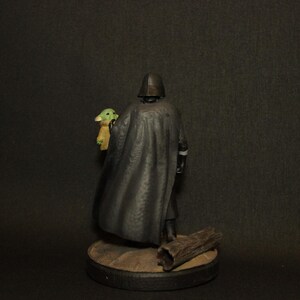 May include: A figurine of a character in a black cape and helmet holding a small green creature with large ears. The figurine is on a round base with a piece of wood. The background is dark.
