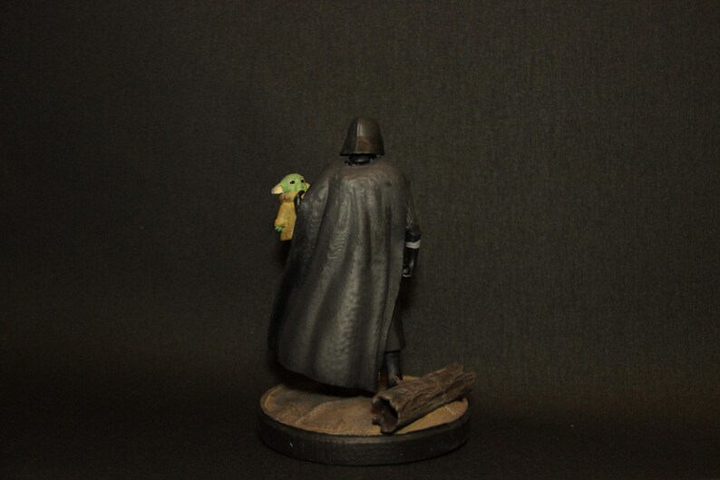 May include: A figurine of a character in a black cape and helmet holding a small green creature with large ears. The figurine is on a round base with a piece of wood. The background is dark.