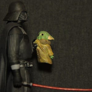 May include: A figurine of Darth Vader holding a Grogu figure, with a red lightsaber in the foreground. Darth Vader is in a black suit and helmet. Grogu is green with large ears and wearing a tan cloak.