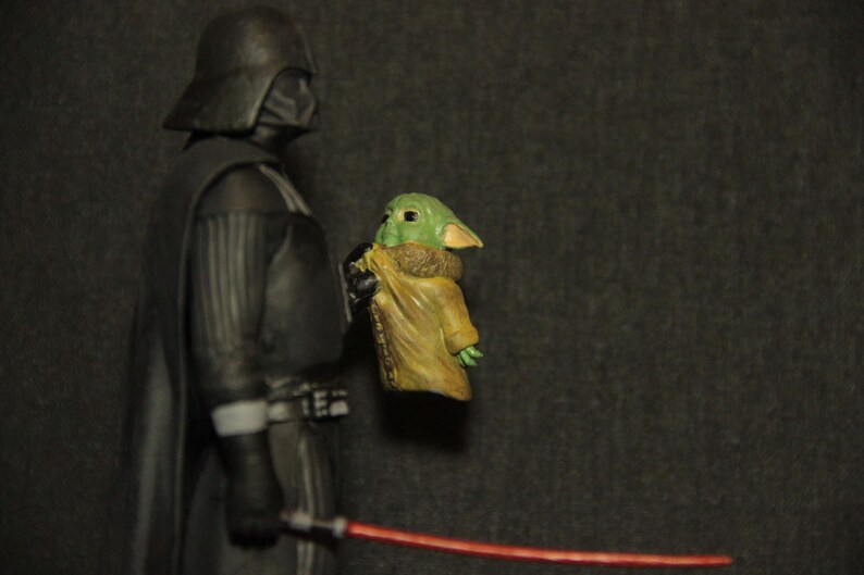 May include: A figurine of Darth Vader holding a Grogu figure, with a red lightsaber in the foreground. Darth Vader is in a black suit and helmet. Grogu is green with large ears and wearing a tan cloak.
