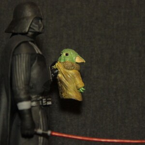 May include: A figurine of Darth Vader holding a Grogu figure, with a red lightsaber in the foreground. Darth Vader is in a black suit and helmet. Grogu is green with large ears and is wrapped in a tan cloth.