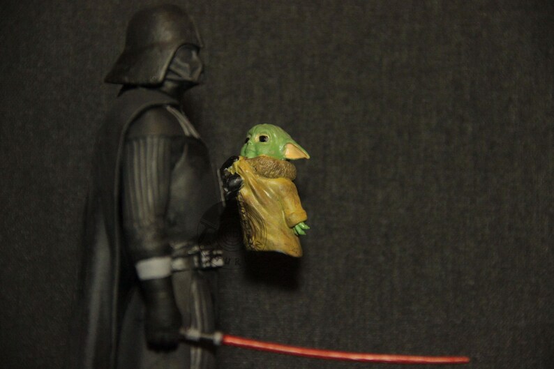 May include: A figurine of Darth Vader holding a Grogu figure, with a red lightsaber in the foreground. Darth Vader is in a black suit and helmet. Grogu is green with large ears and is wrapped in a tan cloth.