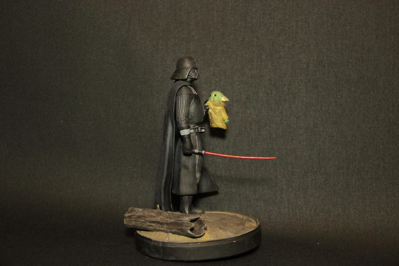 May include: A Star Wars collectible figurine featuring Darth Vader holding a small Grogu figure and a red lightsaber. The figures are on a round base with a piece of wood.