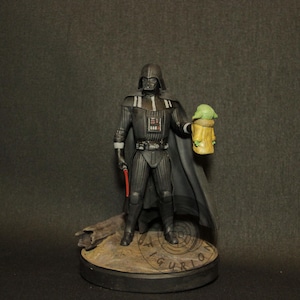 May include: A figurine of Darth Vader holding a red lightsaber and a Grogu figure. The Darth Vader figure is in a black suit with a cape. The Grogu figure is in a tan robe. The base of the figurine is brown.