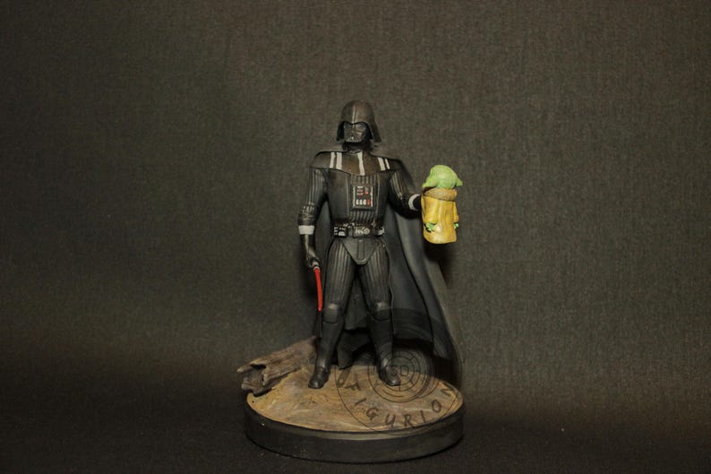 May include: A figurine of Darth Vader holding a red lightsaber and a Grogu figure. The Darth Vader figure is in a black suit with a cape. The Grogu figure is in a tan robe. The base of the figurine is brown.