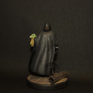 May include: A figurine of Darth Vader holding a Grogu figure. Vader is in a black cape and helmet. Grogu is green with tan clothing. The figures are on a round base with a piece of wood. The background is dark.