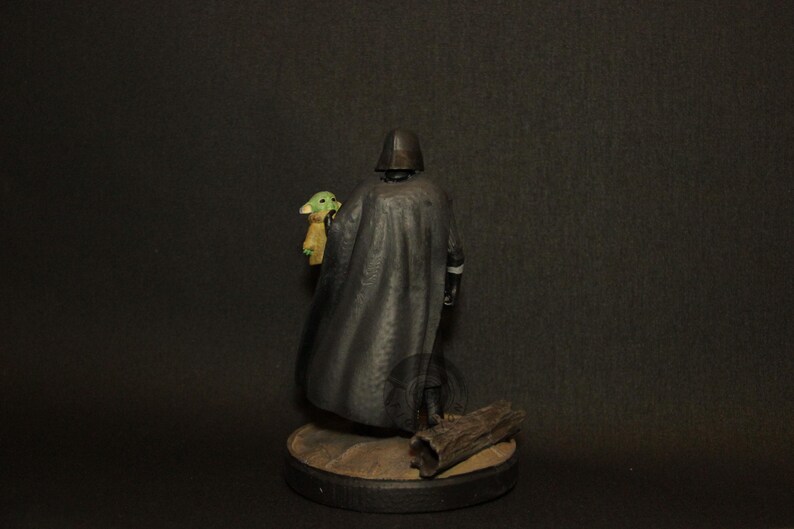 May include: A figurine of Darth Vader holding a Grogu figure. Vader is in a black cape and helmet. Grogu is green with tan clothing. The figures are on a round base with a piece of wood. The background is dark.
