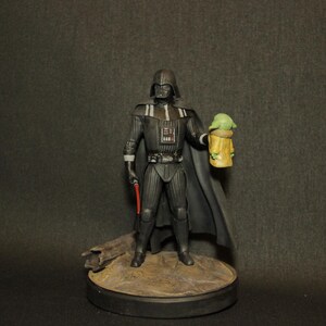 May include: A figurine of Darth Vader holding a red lightsaber and a Grogu figure. The Darth Vader figure is in a black suit with a cape. The Grogu figure is green and yellow. The figures are on a round base.
