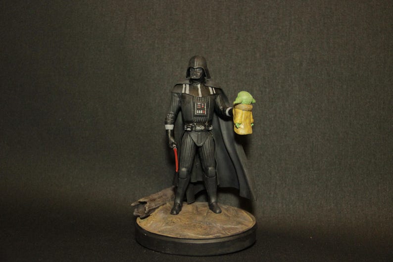 May include: A figurine of Darth Vader holding a red lightsaber and a Grogu figure. The Darth Vader figure is in a black suit with a cape. The Grogu figure is green and yellow. The figures are on a round base.