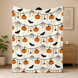 May include: A cream-colored fleece blanket featuring a repeating Halloween pattern of orange pumpkins, black spiderwebs, and black bats hanging from a string of beads and stars. The design includes small orange dots scattered across the fabric.