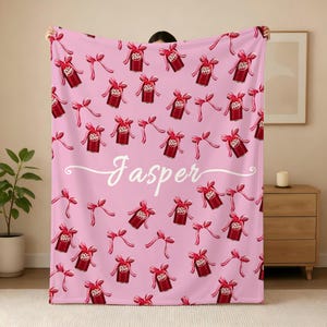 May include: A light pink fleece blanket featuring a repeating pattern of red soda cans tied with pink bows. The name Jasper is printed in white cursive script across the center of the blanket. The design is consistent across the entire fabric surface.
