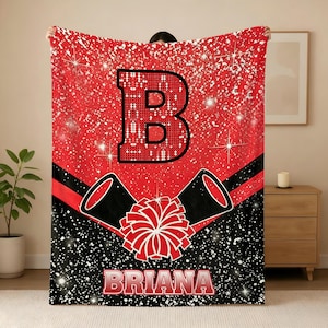 May include: A rectangular throw blanket featuring a red and black color scheme with a glittery effect. The design includes a large letter B, two black megaphones, a red pom-pom, and the name BRIANA printed in white letters with a red outline.