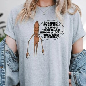 May include: A light gray t-shirt featuring a cartoonish brown creature with long limbs and a speech bubble. The speech bubble contains the text: "APPARENTLY, IT'S NOT LEGAL TO LAUNDER 17,000 DOLLARS THROUGH A LOCALLY OWNED GREEK RESTAURANT."