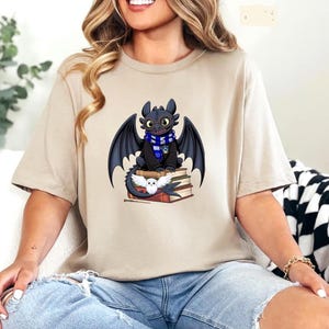 May include: Beige t-shirt featuring a cartoon dragon with black wings, a blue and white scarf, and a black robe, sitting on a stack of books. A white owl is also on the books. The dragon is a popular character from a fantasy movie.
