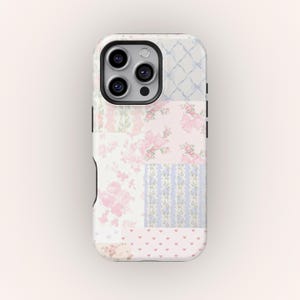 Puede incluir: Una funda de teléfono decorativa con un diseño de patchwork. La funda presenta varios patrones, incluyendo estampados florales, lazos y corazones, en tonos de rosa, azul y blanco. La funda tiene un módulo de cámara negro.