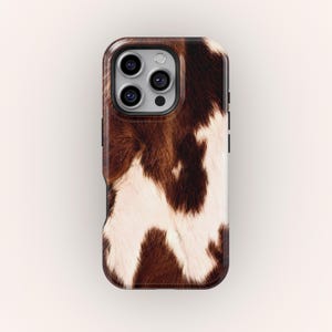 Könnte beinhalten: Smartphone-Hülle mit einem braunen und weißen Kuhfellmuster. Die Hülle hat eine glänzende Oberfläche und einen dunklen Rahmen mit Aussparungen für Kamera und Tasten. Das Design ahmt das natürliche Aussehen von Tierfell nach.