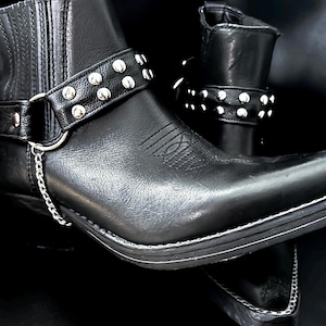 May include: Black leather ankle boots with silver stud detailing on the straps. The boots feature a pointed toe, decorative stitching, and a silver chain. The boots are set against a dark backdrop.