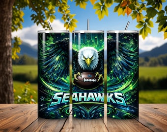 Pode incluir: Três canecas isoladas de aço inoxidável com um design vibrante. O design apresenta uma águia majestosa segurando uma bola de futebol, contra uma paisagem urbana e um fundo de tons verdes e azuis. A palavra "SEAHAWKS" é exibida de forma proeminente.