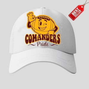 以下が含まれることがあります： 白い野球帽。漫画のフットボールのデザインで、ウィンクして「OK」サインをしており、マルーンとゴールドで「COMANDERS Pride」の文字が書かれています。赤い「BEST SELLER」タグが付いています。