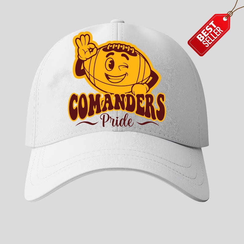 以下が含まれることがあります： 白い野球帽。漫画のフットボールのデザインで、ウィンクして「OK」サインをしており、マルーンとゴールドで「COMANDERS Pride」の文字が書かれています。赤い「BEST SELLER」タグが付いています。