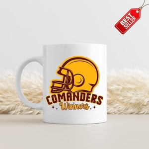 以下が含まれることがあります： 金とマルーンのフットボールヘルメットのグラフィックが特徴の白いセラミックマグ。マグには、マルーンに金の星が付いた「COMANDERS Winners」という文字が書かれています。赤い「BEST SELLER」タグが上から吊り下げられています。