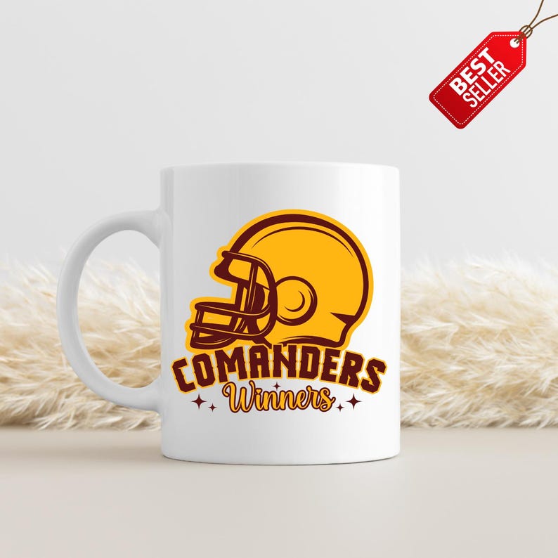 以下が含まれることがあります： 金とマルーンのフットボールヘルメットのグラフィックが特徴の白いセラミックマグ。マグには、マルーンに金の星が付いた「COMANDERS Winners」という文字が書かれています。赤い「BEST SELLER」タグが上から吊り下げられています。