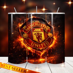Op de afbeelding: Drie roestvrijstalen bekers met het Manchester United-logo. Het logo, een schild met rode en gouden details, is geplaatst tegen een achtergrond van vuur. De bekers hebben een zwarte achtergrond en een rietje.