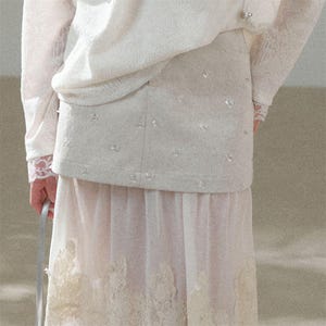 Puede incluir: Un conjunto color crema con una parte superior de manga larga con detalles de encaje y una falda. La falda es de color rosa claro con una superposición transparente y ribete de encaje en el dobladillo. El conjunto está adornado con pequeños adornos brillantes.
