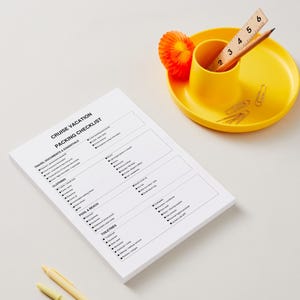Könnte beinhalten: Ein weißer Papierblock mit der Aufschrift Cruise Vacation Packing Checklist in schwarzer Schrift, begleitet von einem gelben Schreibtisch-Organizer mit einem Holzlineal bis 15 cm, einem Bleistift und Büroklammern sowie zwei weiteren Stiften.