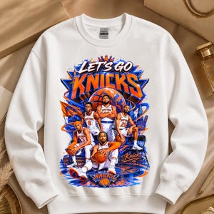 May include: White sweatshirt featuring a graphic of the New York Knicks basketball team. The design includes the words "Let's Go Knicks" in orange and blue, along with illustrations of players and a basketball court.