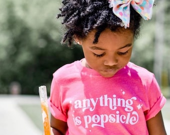 May include: A bright pink short-sleeved t-shirt with the white text "anything is popsicle" printed on the front, accompanied by small star accents. The shirt is paired with dark blue shorts and a large, sheer, multi-colored polka dot hair bow.