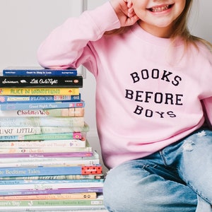 May include: A young girl wearing a pink sweatshirt with the text "BOOKS BEFORE BOYS" in black lettering. She is sitting on a carpeted floor with a stack of children's books in front of her.