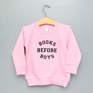 May include: Pink sweatshirt with black text that reads "BOOKS BEFORE BOYS".
