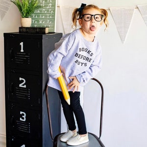 May include: A young girl wearing a grey sweatshirt with the text "BOOKS BEFORE BOYS" in black lettering. She is standing on a metal chair holding a large yellow pencil. She is wearing black pants and white slip-on shoes.