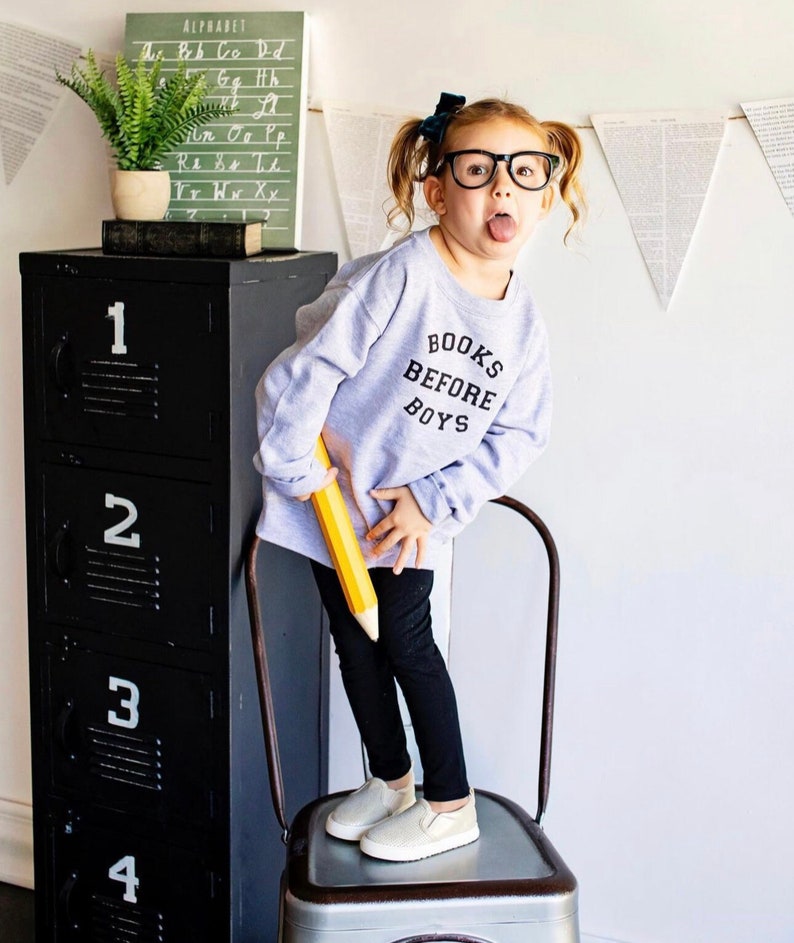 May include: A young girl wearing a grey sweatshirt with the text "BOOKS BEFORE BOYS" in black lettering. She is standing on a metal chair holding a large yellow pencil. She is wearing black pants and white slip-on shoes.