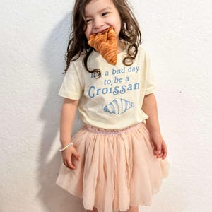 May include: A young person wearing a white t-shirt with the text "It's a bad day to, be a Croissant" and a blue illustration of a croissant. The person is also wearing a pink tulle skirt and pink sandals with yellow sunflower designs.