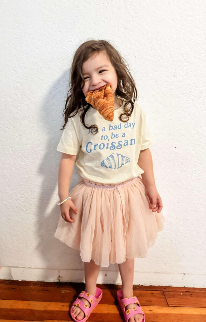 May include: A young person wearing a white t-shirt with the text "It's a bad day to, be a Croissant" and a blue illustration of a croissant. The person is also wearing a pink tulle skirt and pink sandals with yellow sunflower designs.