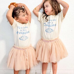 May include: Two young girls wearing white t-shirts with a croissant graphic and the text "It's a good day to be a croissant" and "Good day to be a croissant". They are wearing pink tulle skirts and holding croissants above their heads.