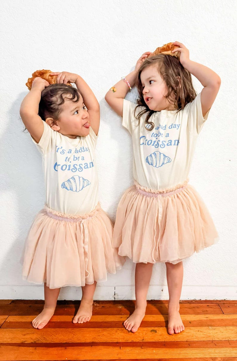 May include: Two young girls wearing white t-shirts with a croissant graphic and the text "It's a good day to be a croissant" and "Good day to be a croissant". They are wearing pink tulle skirts and holding croissants above their heads.